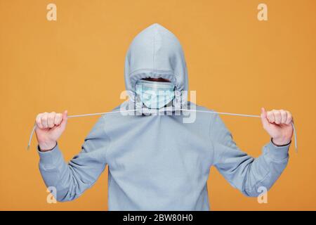 Contemporary young man in hoodie and protective mask stretching cords while standing in front of camera over yellow background Stock Photo