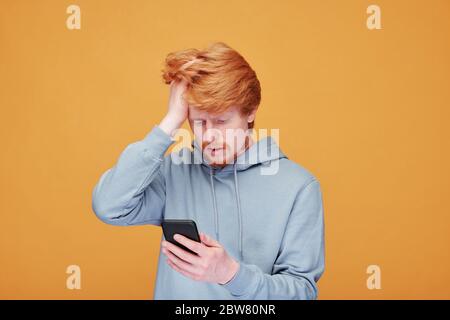 Worried young man in casualwear touching head while reading online news on screen of smartphone against yellow background Stock Photo