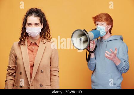 Young man in protective mask shouting in megaphone to female in formalwear standing in front of camera on yellow background Stock Photo