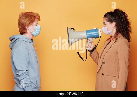 Side view of young brunette woman in protective mask speaknig in megaphone while standing in front of guy in blue hoodie Stock Photo