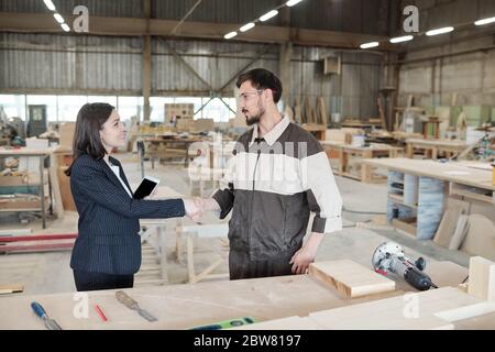 Pretty young female manager or business partner shaking hand of worker of furniture factory after negotiation in large warehouse Stock Photo