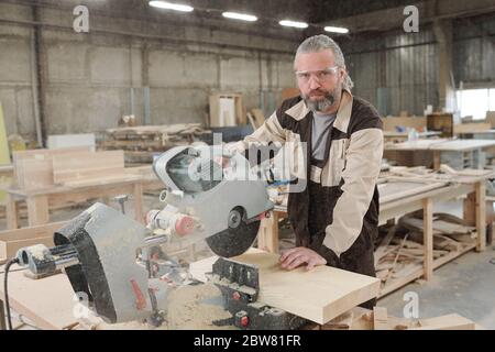 Confident aged professional in eyeglasses and workwear standing by workplace while cutting thick wooden board with electric saw Stock Photo