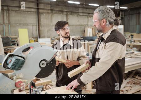 Mature factory worker in workwear looking at young colleague while consulting him about how to use electric circle saw Stock Photo