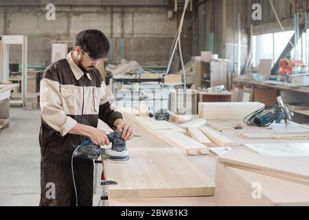 Young male worker in workwear and protective eyeglasses using grinding machine for processing surface of wooden board by workbench Stock Photo