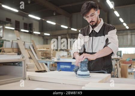 Young man in workwear and protective eyeglasses bending over workbench while processing surface of wooden board Stock Photo