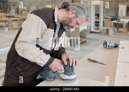 Mature worker of furniture producing factory bending over wooden board while using grinder for smoothing surface of workpiece Stock Photo