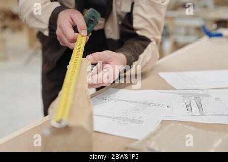 Hands of contemporary worker of factory holding wooden workpiece and measuring its length by workplace with sketches of this detail Stock Photo