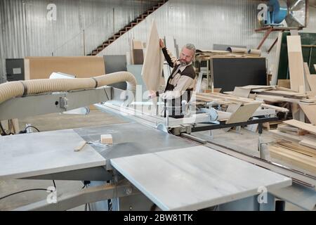 Senior worker of furniture producing factory in uniform putting large heavy wooden board on industrial machine before processing it Stock Photo