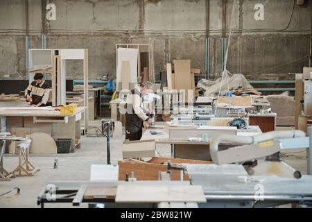 Two workers of large contemporary furniture producing factory standing by workbenches in workshop and drilling wooden workpieces Stock Photo