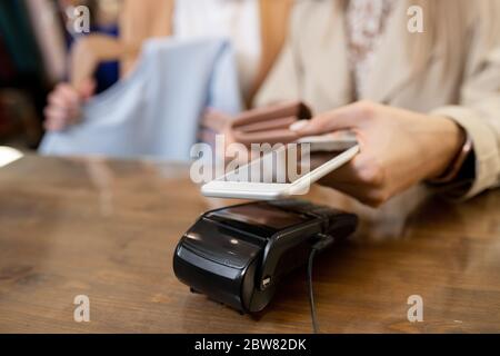 Hand of young female shopaholic holding smartphone over payment machine on counter while buying new clothes in boutique Stock Photo