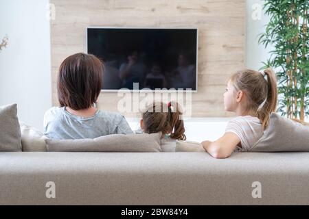 happy mother with her two cute daughters watching tv at home. Happy family relaxing on the coach. Stock Photo
