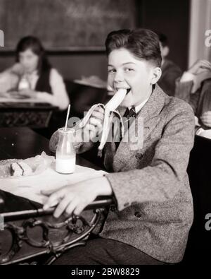 1920s BOY STUDENT LOOKING AT CAMERA EATING SNACK AT HIS SCHOOL DESK A BANANA BOTTLE OF MILK WITH STRAW CAKE SLICE IN WAXED PAPER - f4151 HAR001 HARS NOSTALGIA OLD FASHION 1 JUVENILE ELEMENTARY STYLE COMMUNICATION SNACK PLEASED JOY LIFESTYLE SATISFACTION DAIRY COPY SPACE HALF-LENGTH BANANA MALES B&W EYE CONTACT SCHOOLS SUIT AND TIE GRADE HAPPINESS CHEERFUL BEVERAGE HIS FLUID NUTRITION PRIMARY SMILES CONSUME CONSUMING JOYFUL NOURISHMENT STYLISH WAXED COOPERATION GRADE SCHOOL GROWTH JUVENILES PROTEIN BLACK AND WHITE CAUCASIAN ETHNICITY HAR001 OLD FASHIONED Stock Photo