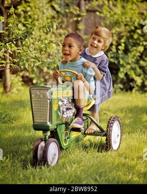 1960s AFRICAN-AMERICAN BOY DRIVING A TOY PEDDLE TRACTOR IN BACKYARD BAREFOOT CAUCASIAN GIRL HITCHING A RIDE ON BACK FOOT STEP - kj5460 PHT001 HARS 1 FOOT SILLY JUVENILE DIVERSE BLOND FRIEND COMIC TEAMWORK RIDE DIFFERENT JOY LIFESTYLE FEMALES TRACTOR RURAL HEALTHINESS HOME LIFE COPY SPACE FRIENDSHIP FULL-LENGTH HALF-LENGTH INSPIRATION MALES HUMOROUS HAPPINESS LEISURE AFRICAN-AMERICANS AFRICAN-AMERICAN AND LOW ANGLE BLACK ETHNICITY COMICAL A IN ON BAREFOOT CONNECTION PEDDLE CONCEPTUAL COMEDY FRIENDLY HITCHING VARIOUS VARIED COOPERATION JUVENILES TOGETHERNESS CAUCASIAN ETHNICITY OLD FASHIONED Stock Photo