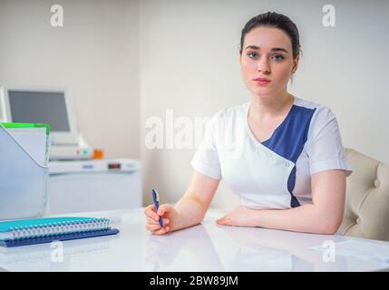 Female young doctor sitting at her desk holding pen and looking in camera Stock Photo