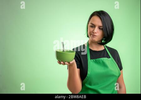 Female cook with green apron and black t-shirt, she looks at the bowl she is holding in her hand isolated on green background Stock Photo