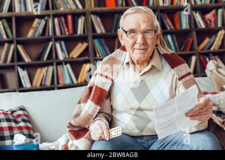 Senior man alone sitting at library sick covered with plaid holding pills and prescribing information thoughtful Stock Photo