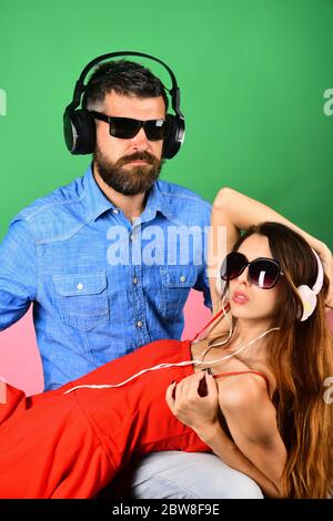 Relax, party and music concept. Music fans with serious faces enjoy music. Man with beard holds girl lying on green and pink background. Couple in love wears headphones and sunglasses. Stock Photo