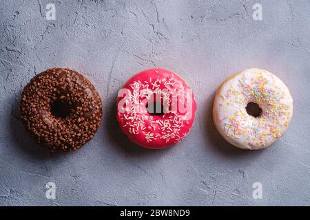 Three donuts in row, chocolate, pink and vanilla donut with sprinkles on, sweet glazed dessert food on concrete textured background, top view Stock Photo