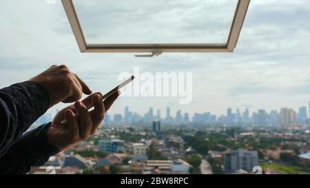 A man types a message on a smartphone against the background of an open window and a panorama of the city of Jakarta. Hands close up. Stock Photo