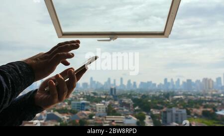A man types a message on a smartphone against the background of an open window and a panorama of the city of Jakarta. Hands close up. Stock Photo