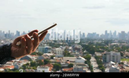 A man holds a smartphone on the background of the panorama of the city of Jakarta Stock Photo
