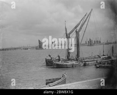 A Thames estuary view . The dock cranes can be seen in the distance . In the foreground two sailing barges are are moored on the side of the river . 1939 Stock Photo