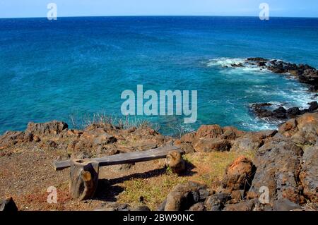 Horizon stretches out in deep blue aqua along the rocky stretch of shoreline at Lapakahi State Historical Park.  A single rustic log-hewn bench sits o Stock Photo