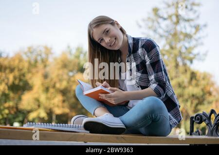 University student studying, learning language, taking notes, exam preparation sitting on bench. Portrait of young happy woman working in park Stock Photo