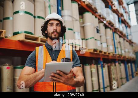 Young caucasian male worker doing stocktaking of product in cardboard box on shelves in warehouse by using digital tablet Stock Photo