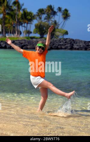 Adult female celebrates retirement by kicking and splashing on the beach on the Big Island of Hawaii.  She is wearing a sunvisor and sunglasses.  Coul Stock Photo
