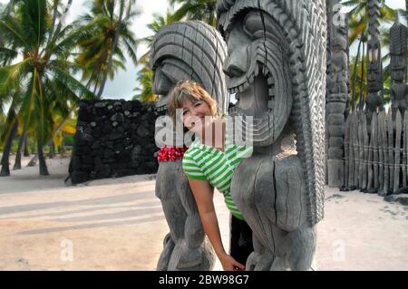 Woman tourist leans between two tiki at the Puuhonua o Honaunau National Historical Park on the Big Island of Hawaii.  She is smiling and happy as she Stock Photo