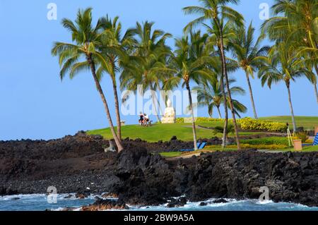 Couple relax on a bench beneath palm trees at a resort on the Big Island of Hawaii.  Ocean waves splash against the lava rock beach. Stock Photo