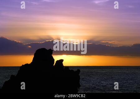 Rising sun colors the clouds and sky purple and glows golden and orange.  Sea stacks, which are lava rocks jutting up from the ocean, are silhouetted Stock Photo