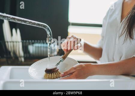 Young woman washes dishes with wooden brush with natural bristles at window in kitchen. Zero waste concept Stock Photo