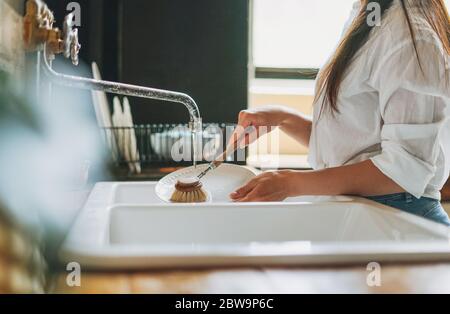 Young woman washes dishes with wooden brush with natural bristles at window in kitchen. Zero waste concept Stock Photo