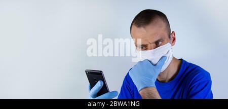 Young doctor in a protective mask and blue clothes on a blue background. looking at a mobile phone in his hand.copyspace for text. Stock Photo