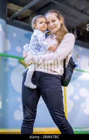 A cute young Caucasian mother holds a baby daughter in her arms, looks at the camera and smiles happily and laughs. vertical photo, close-up, soft