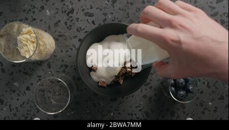 top view man hands preparing breakfast with chocolate granola, almond flakes and blueberry in black bowl on terrazzo surface Stock Photo