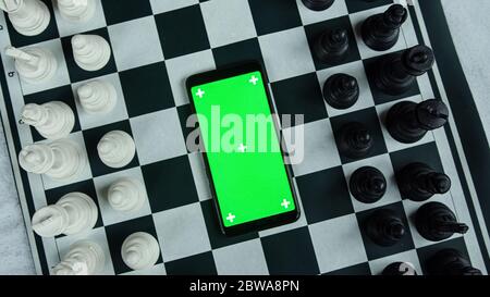 Black and white chess pieces and a smart green screen phone in the middle. Chess application concept Stock Photo