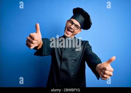 Young handsome chef man wearing cooker uniform and hat over isolated blue background approving doing positive gesture with hand, thumbs up smiling and Stock Photo
