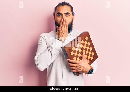 Young arab man holding chess covering mouth with hand, shocked and afraid for mistake. surprised expression Stock Photo