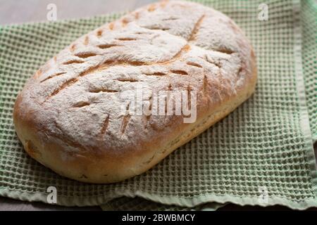 Homemade artisan sourdough bread loaf with artistic leaf pattern scoring and a wavy line in the middle on a green kitchen towel on wooden surface. Stock Photo