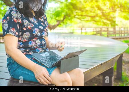 Woman wears protective mask using spray alcohol sanitizer cleaning a laptop to prevent the virus and bacterias in garden. Quarantine for coronavirus ( Stock Photo