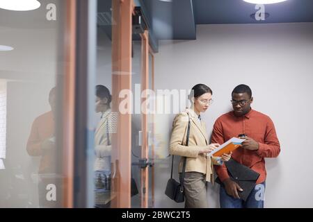 Waist up portrait of two contemporary office workers discussing documents while leaning against wall in hall, copy space Stock Photo