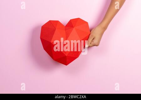 Hand holding red polygonal paper heart shape on light pink background Stock Photo