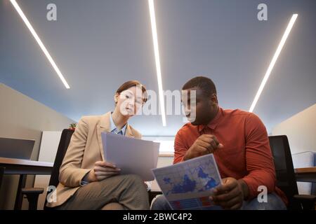 Low angle portrait of two contemporary business people discussing documents during meeting while sitting on chairs in futuristic office interior, copy Stock Photo