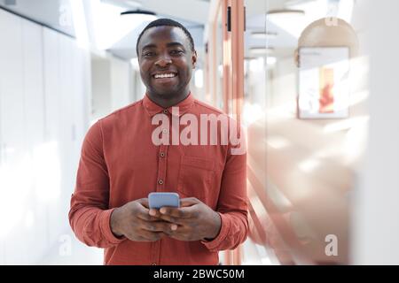 Waist up portrait of contemporary African-American man holding smartphone and smiling at camera while standing in futuristic office interior, copy spa Stock Photo