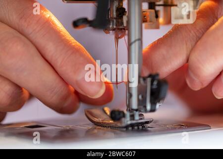elder woman fingers inserting a red thread into a sewing machine's needle, close-up Stock Photo