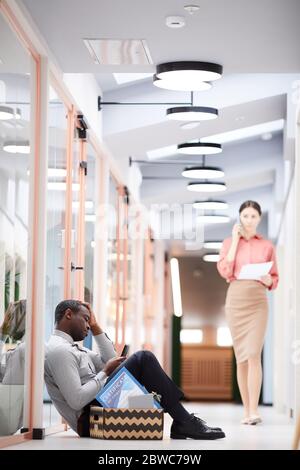 Vertical side view at young African man sitting with box of belongings outside office after being fired, copy space Stock Photo