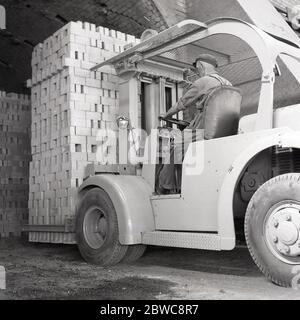 1950s, historical, male operator sitting at the wheel of a forklift truck of the era moving a heavy load of newly-produced London bricks, Stewartby, Bedfordshire, England, UK. Battery powered forklifts were open cockpit industrial trucks used to move materials short distances. Stock Photo